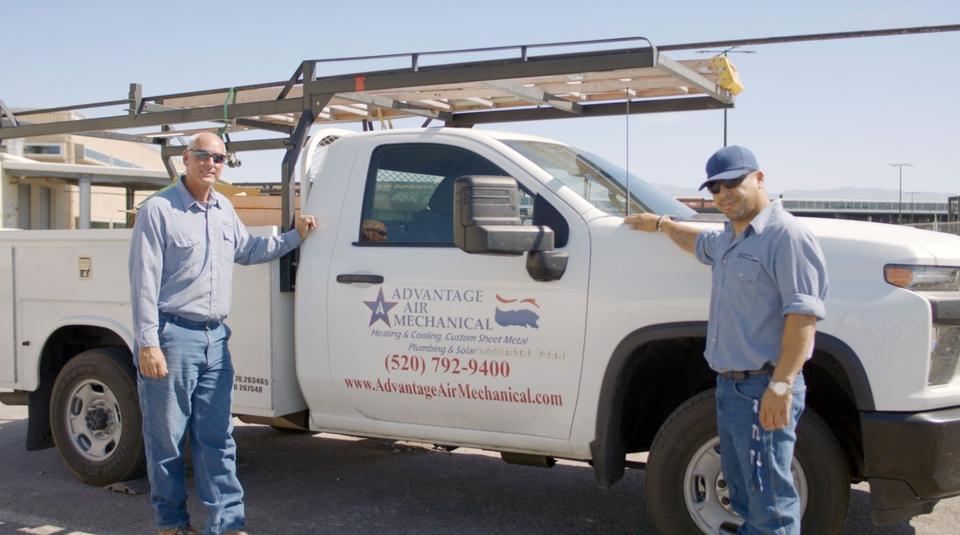 Two uniformed Advantage Air techs leaning against a parked branded service truck, smiling at the camera.