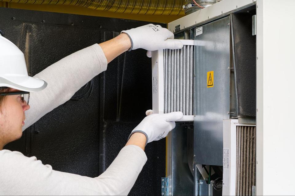 An HVAC tech wearing a white long sleeve shirt, white hard hat, and white gloves, replacing a furnace filter