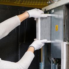 An HVAC tech wearing a white long sleeve shirt, white hard hat, and white gloves, replacing a furnace filter