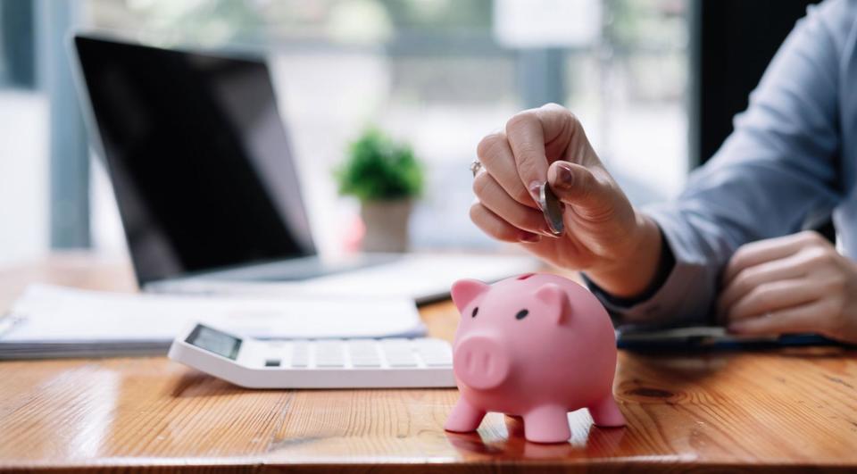 Person putting coin into a piggy bank on their desk with a white calculator and opened laptop nearby