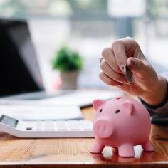 Person putting coin into a piggy bank on their desk with a white calculator and opened laptop nearby