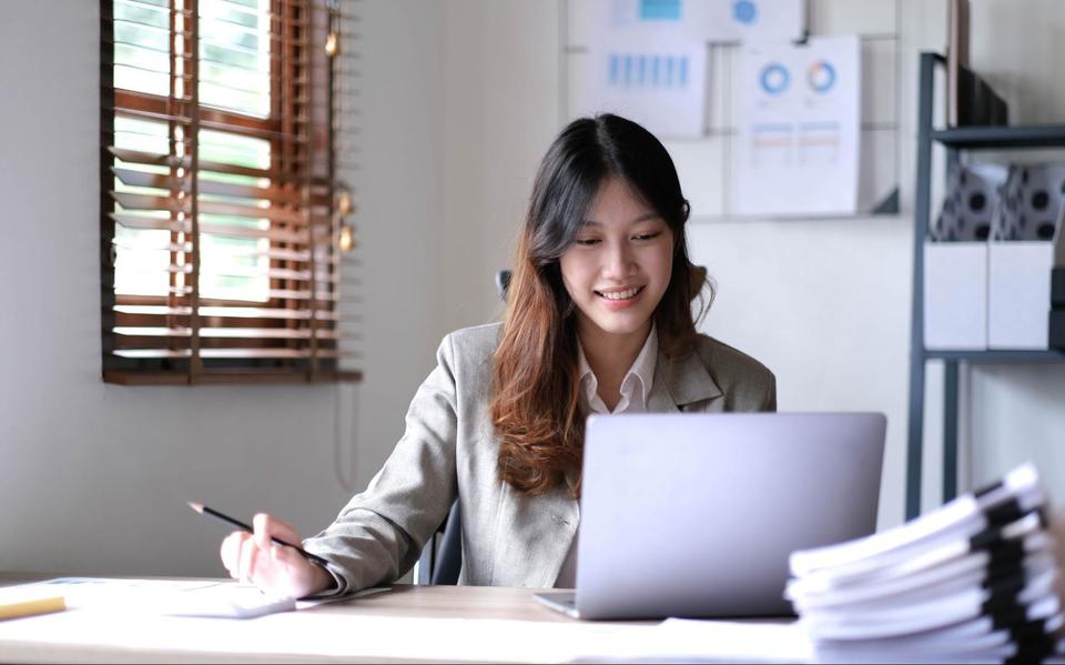 Female accountant in an office, sitting at her desk, smiling at her opened laptop, holding a pencil in her right hand.