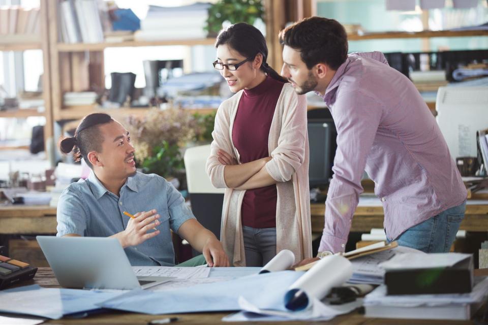 Three business employees, one sitting, and two standing, talking together in an office space