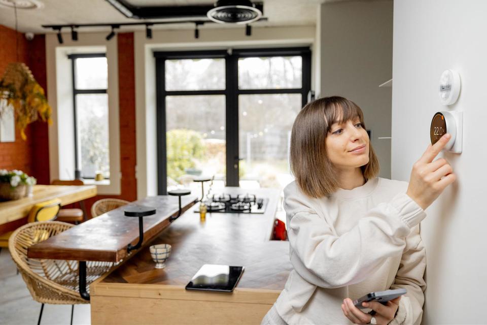 A woman, with short brunette hair, wearing a cream colored sweater, leaning against her white wall, using her right hand to adjust her smart thermostat near the kitchen, while holding her cell phone in her left hand.