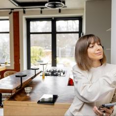 A woman, with short brunette hair, wearing a cream colored sweater, leaning against her white wall, using her right hand to adjust her smart thermostat near the kitchen, while holding her cell phone in her left hand.