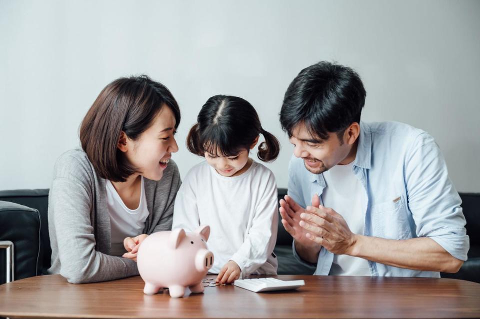 Parents encouraging their daughter to place her coins in a pink piggy bank in front of her, with a white calculator nearby as well.