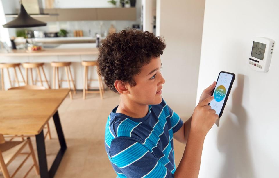 A kid with curly dark brown hair, wearing a blue striped shirt, standing in front of a thermostat on a white wall and adjusting the thermostat from his smart app on his phone.