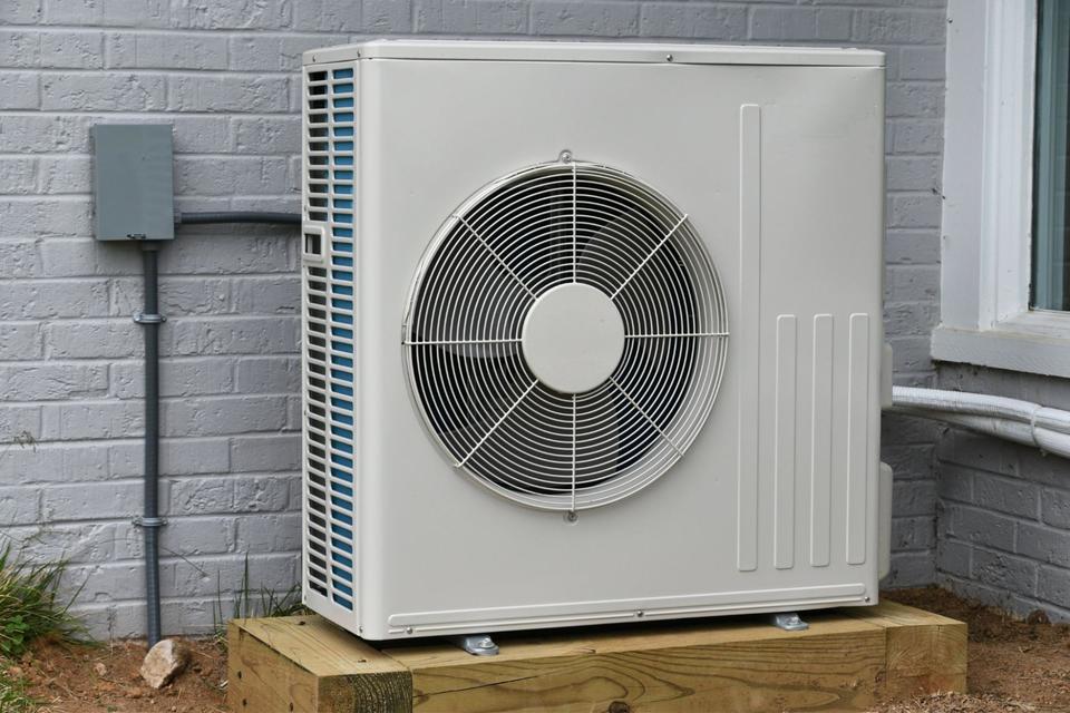 A beige outdoor heat pump unit affixed on a wood block next to a gray block exterior wall.