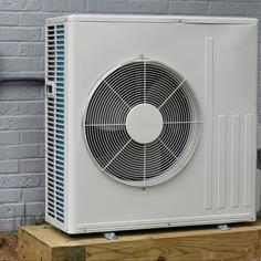 A beige outdoor heat pump unit affixed on a wood block next to a gray block exterior wall.