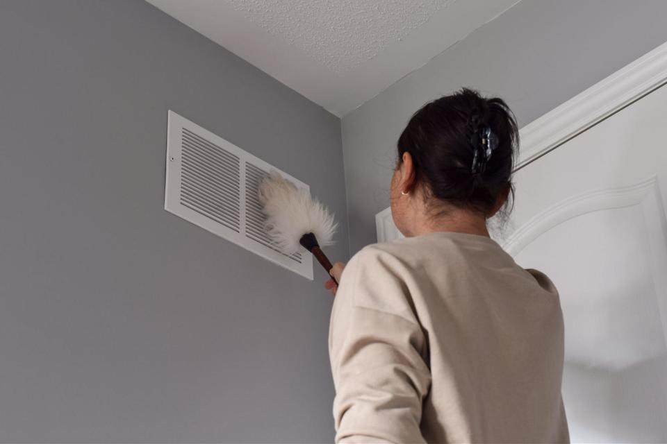 Woman with her up in a clip, wearing a tan sweater and dusting her white vent affixed to a light gray wall.