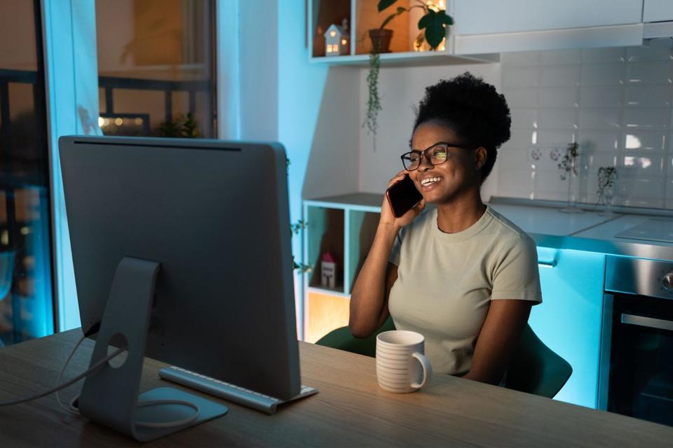 Woman sitting at a table in her kitchen, smiling while holding her cell phone up to her right ear with a white coffee mug in front of her and looking at her computer screen.