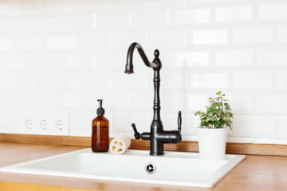 A white kitchen sink with a black faucet, installed in a wooden countertop, with a white tiled backsplash behind it.