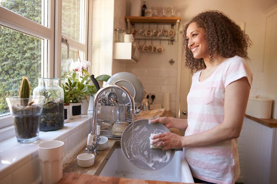 A woman wearing a pink and white stripe shirt, standing at her kitchen counter, smiling while looking out the window and washing dishes in her farmhouse sink.
