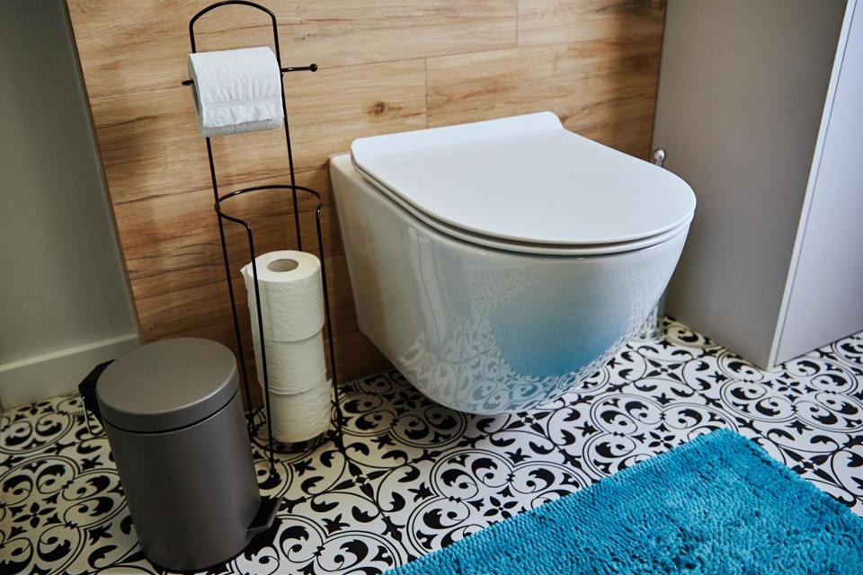 A toilet affixed to a wood tiled wall with a small metal trashcan and a toilet paper holder to the right of it and with a black and white tiled floor underneath it with a bright blue rug in front of it.