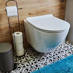 A toilet affixed to a wood tiled wall with a small metal trashcan and a toilet paper holder to the right of it and with a black and white tiled floor underneath it with a bright blue rug in front of it.