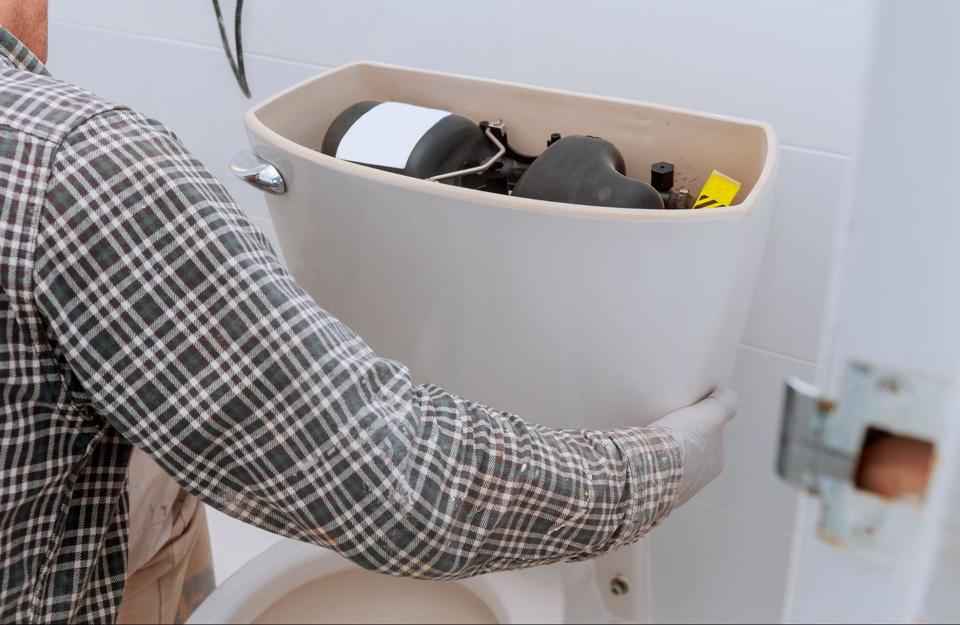 A man wearing a plaid long sleeve shirt is kneeling down next to a toilet with it's tank lid off, showing the pump and inside of tank.