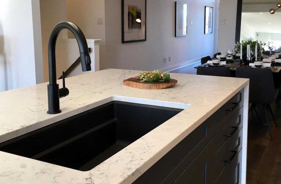A black kitchen faucet installed with a one basin black sink in a white quartz countertop on a kitchen island with black cabinetry.