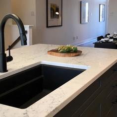 A black kitchen faucet installed with a one basin black sink in a white quartz countertop on a kitchen island with black cabinetry.