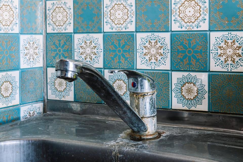 Close up of a silver kitchen faucet and sink that has hard water stains and rust that is installed in front of a white and blue checkered tile backsplash.