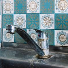 Close up of a silver kitchen faucet and sink that has hard water stains and rust that is installed in front of a white and blue checkered tile backsplash.