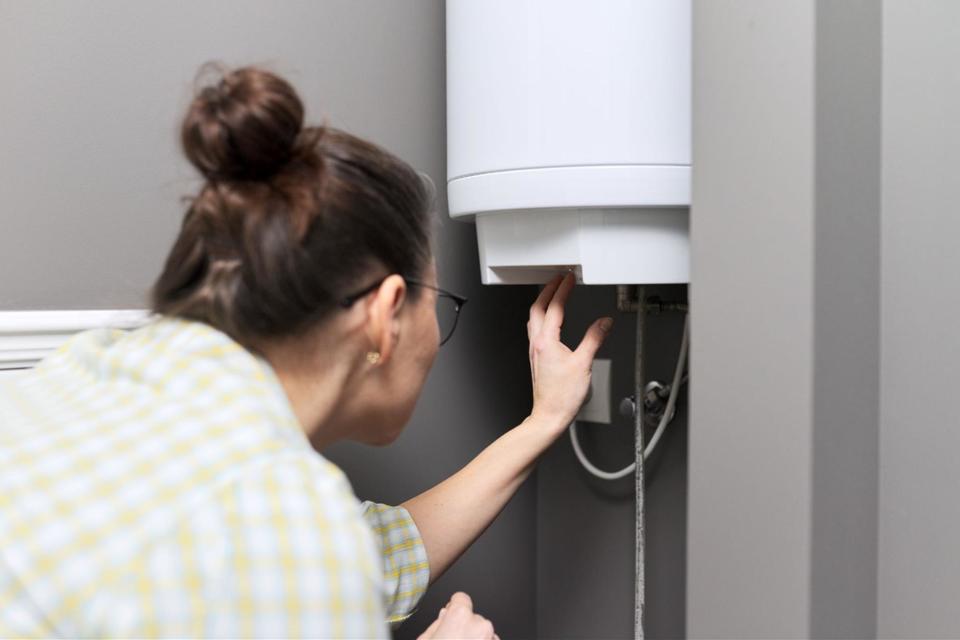 A woman wearing her brunette hair in a bun, black glasses, and a yellow and white plaid shirt checking a part on her water heater.