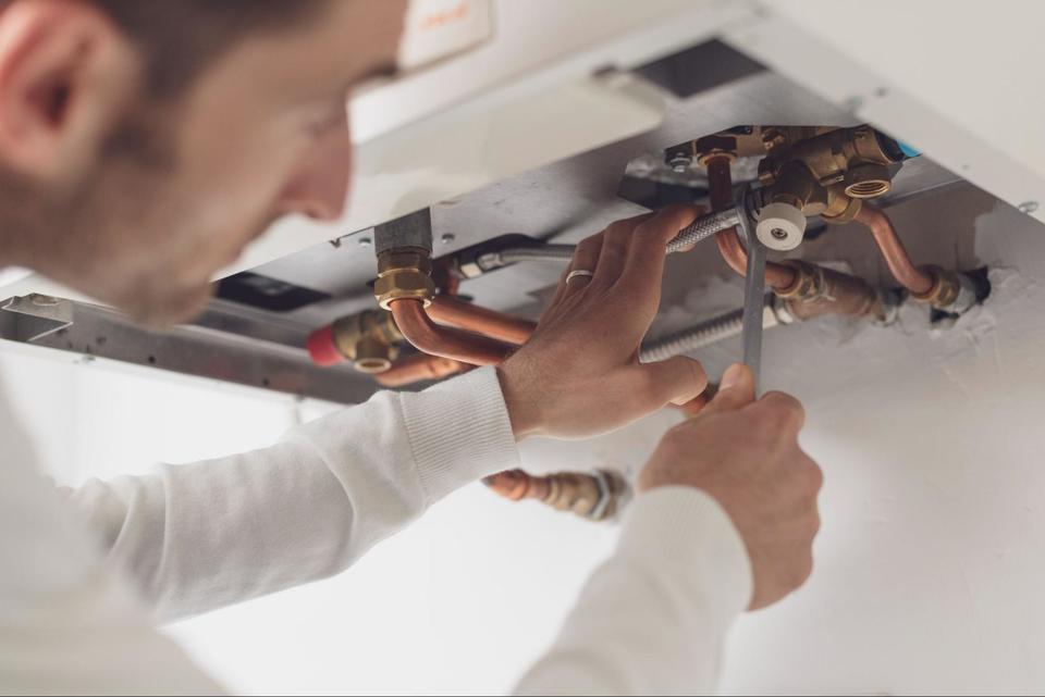 Close up of a tech wearing a white long sleeve shirt, using a wrench to tighten a pipe fitting at the bottom of a water heater.