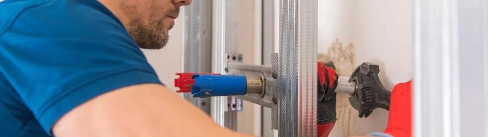 A male tech wearing a blue short sleeved shirt and bright red work gloves is wrenching a pipe on a water heater.