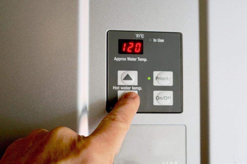 Closeup of a square, dark gray control panel on the front of a water heater with a pointer finger pressing the down arrow on the hot water temp setting.