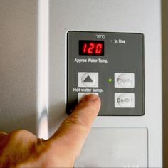 Closeup of a square, dark gray control panel on the front of a water heater with a pointer finger pressing the down arrow on the hot water temp setting.