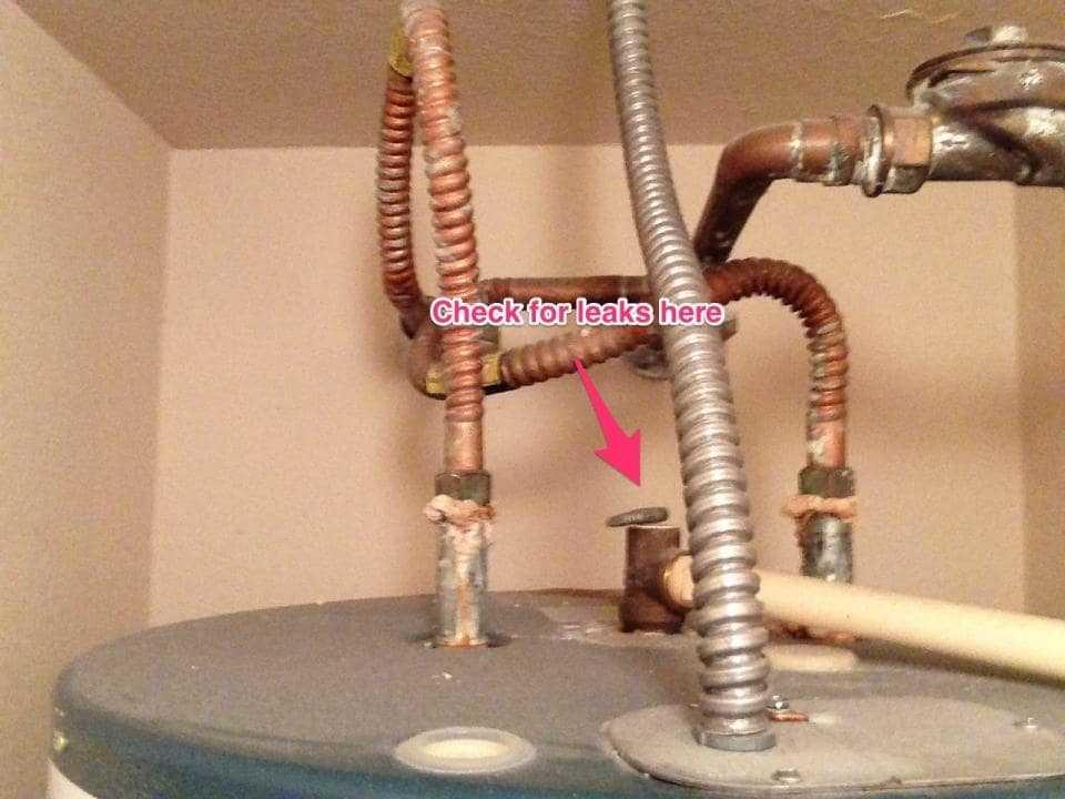 Closeup of the piping on top of a tank water heater with a red arrow pointing to a pipe and with the words "Check for leaks here".