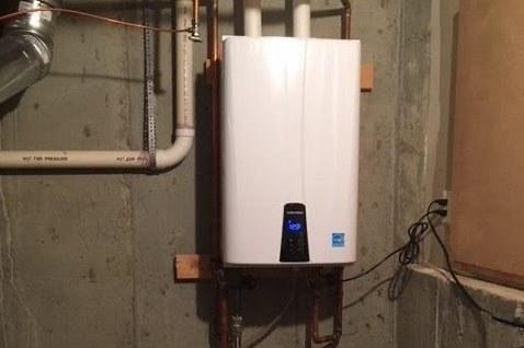 A white tankless water heater affixed to an interior concrete wall surrounded by other various piping.