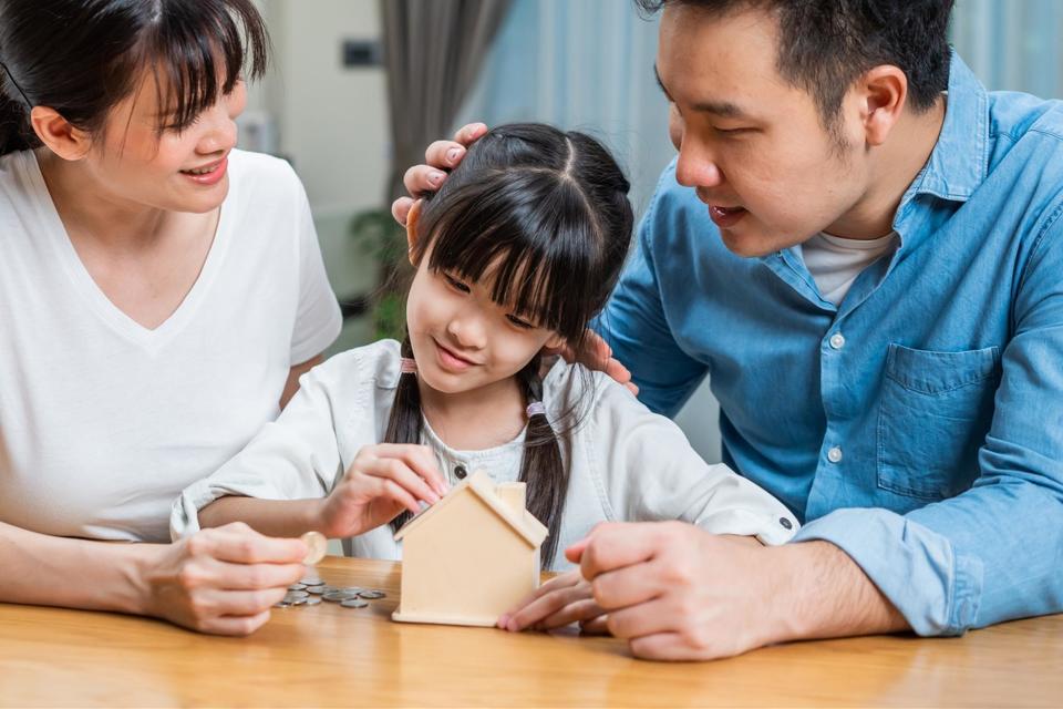 A mom and dad sitting at a wood kitchen table smiling at their daughter while she puts coins in a house shaped piggy bank