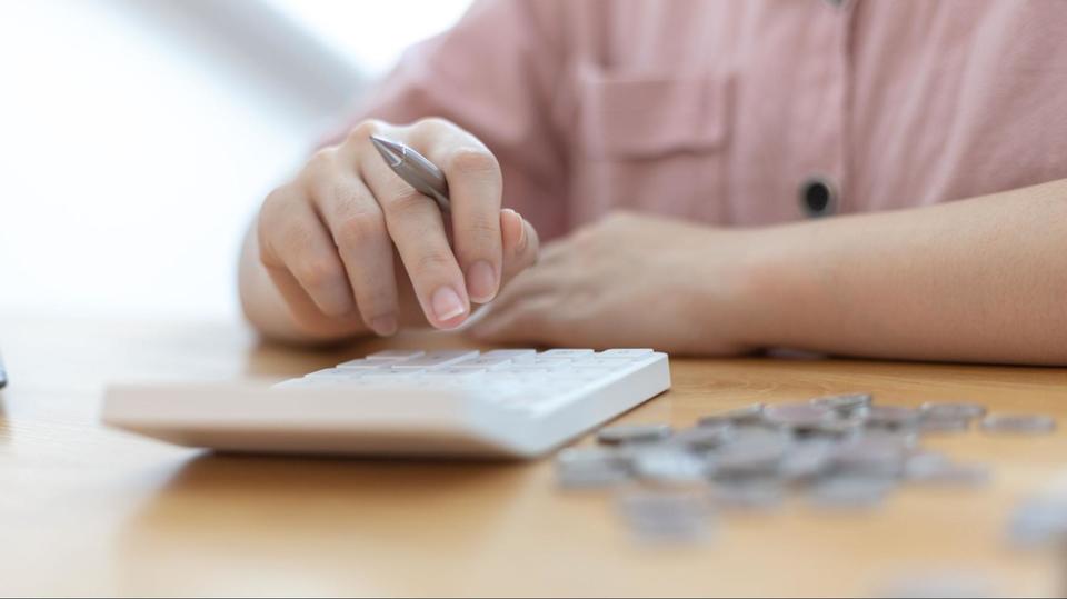 Person using a white calculator sitting on a wooden table with silver coins in a pile next to the calculator