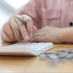 Person using a white calculator sitting on a wooden table with silver coins in a pile next to the calculator