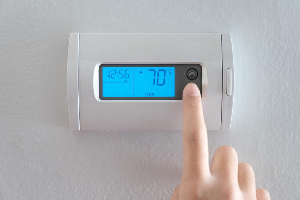 A finger pressing the down button on a white thermostat affixed to a white wall.