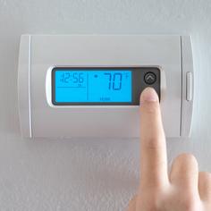 A finger pressing the down button on a white thermostat affixed to a white wall.