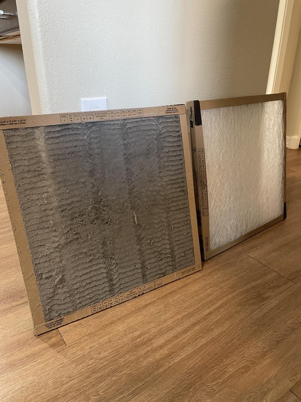 One square dirty air filter sitting next to a clean square air filter on a wood plank floor.