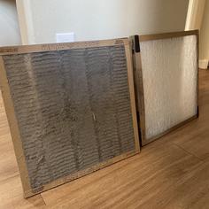 One square dirty air filter sitting next to a clean square air filter on a wood plank floor.
