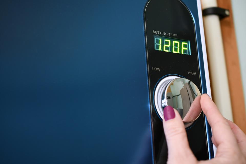 A woman with maroon painted nails adjusting the temperature on her tankless water heater using a silver knob.