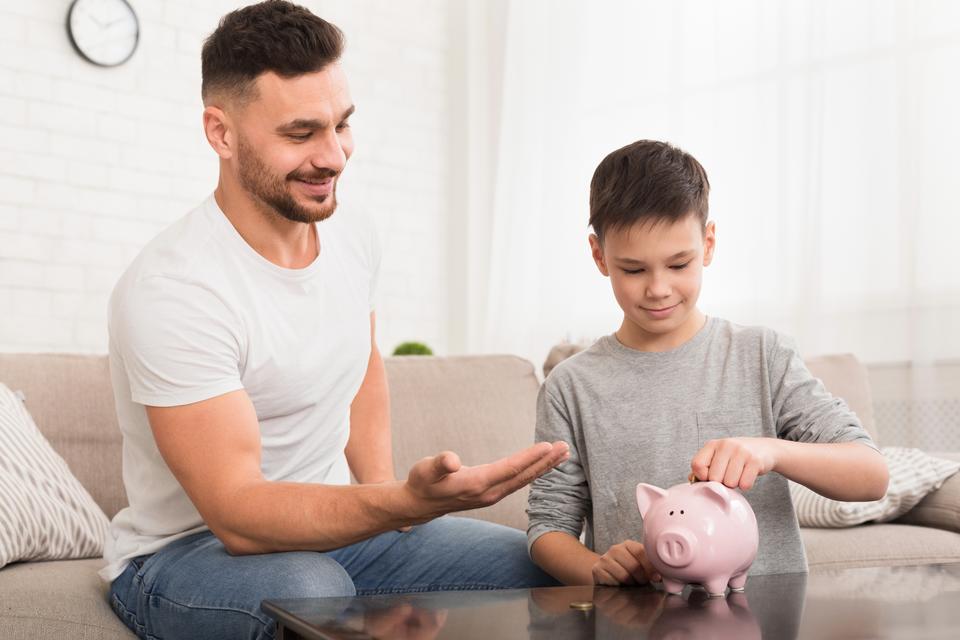 A father sitting next to his son, with his right hand out and holding coins while his son puts them into his pink piggy bank sitting on the coffee table.